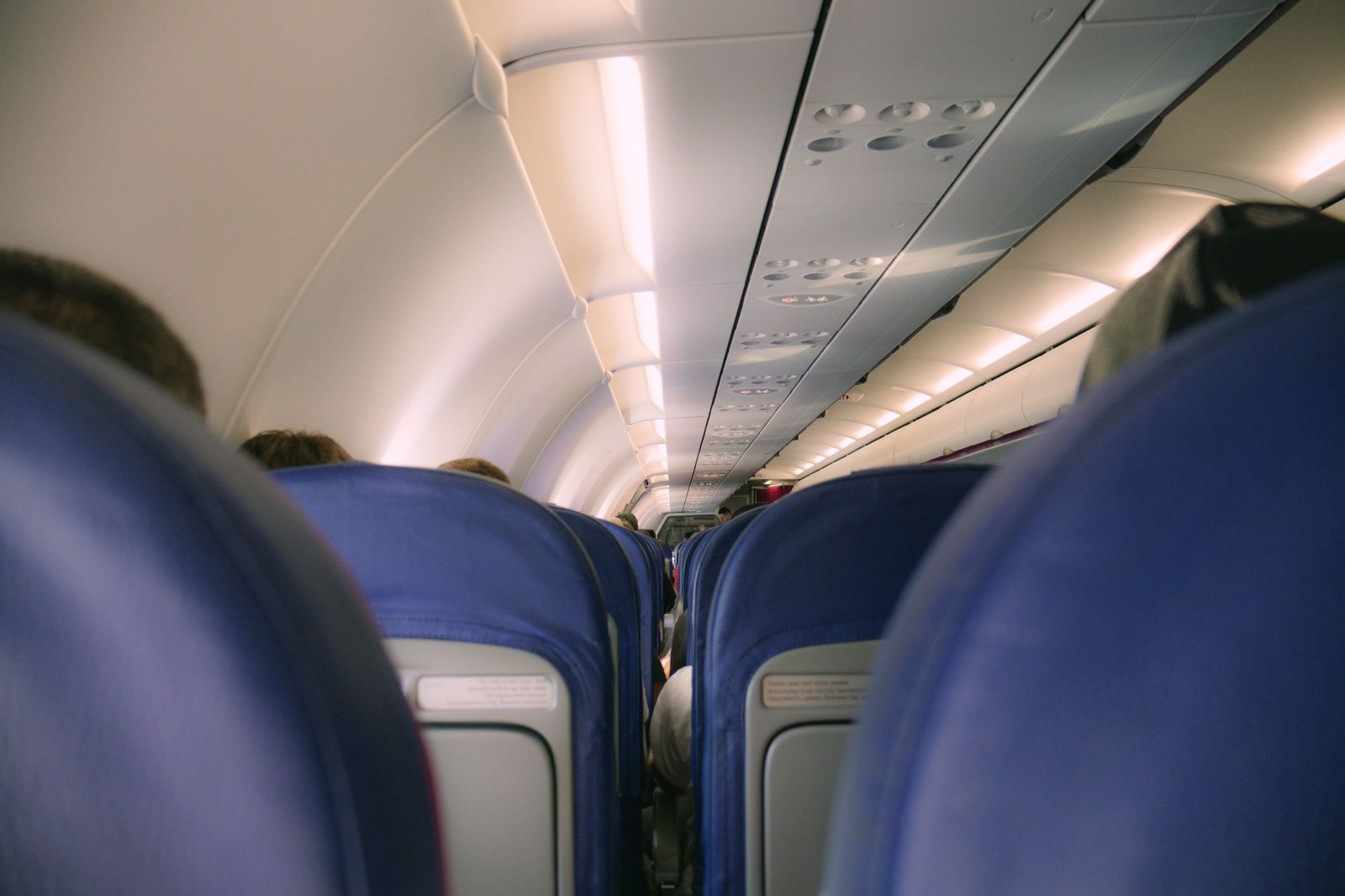 View down the aisle of a narrow-body aircraft, rows of blue economy seats on both sides with seated passengers, overhead lockers lit above.
